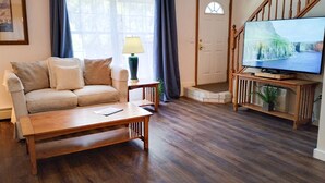 A living room with a wooden floor, a beige sofa, a coffee table, a television on a wooden stand, and a door with a white frame. - Cozy Dog Friendly House with Deck, Firepit & Fast WiFi in Heart of Bar Harbor (Bar Harbor)