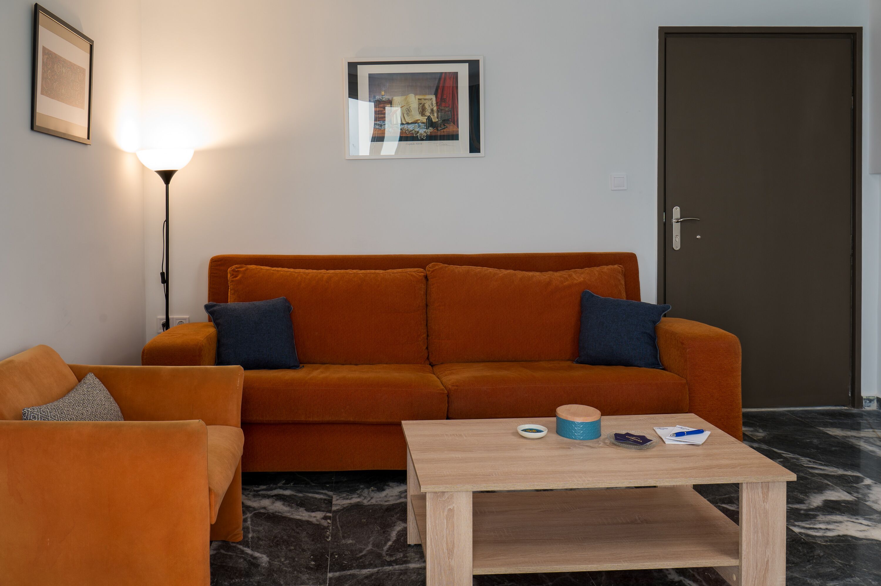 Una habitación con un sofá naranja, una mesa de centro de madera, un cuadro en la pared y una puerta.