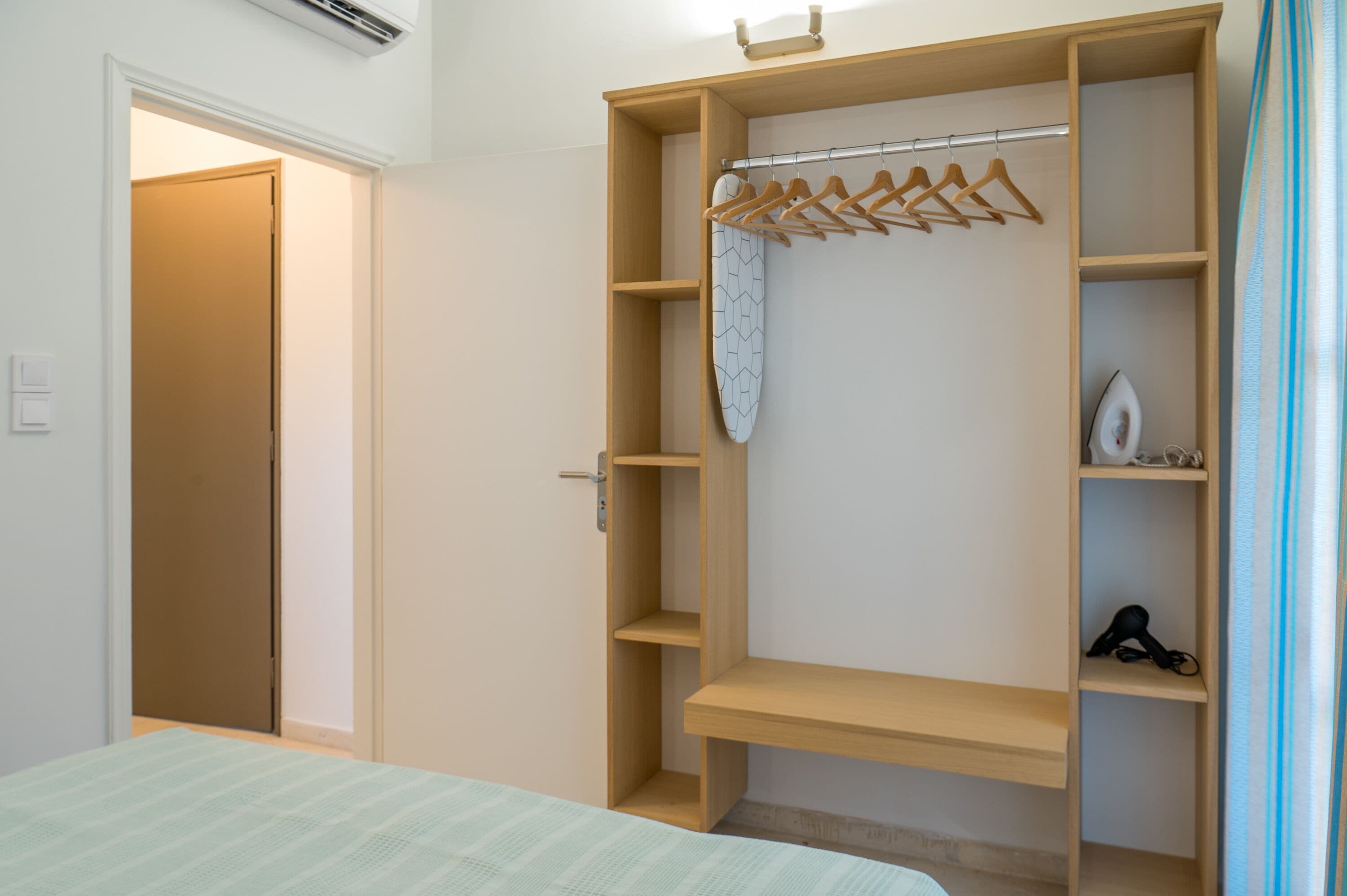 A bedroom with a bed, a wooden wardrobe with hangers, and a shelf with a hairdryer.