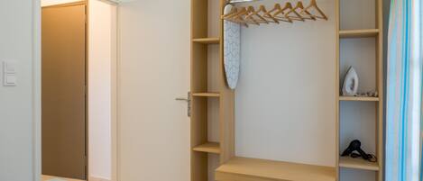 A bedroom with a bed, a wooden wardrobe with hangers, and a shelf with a hairdryer.