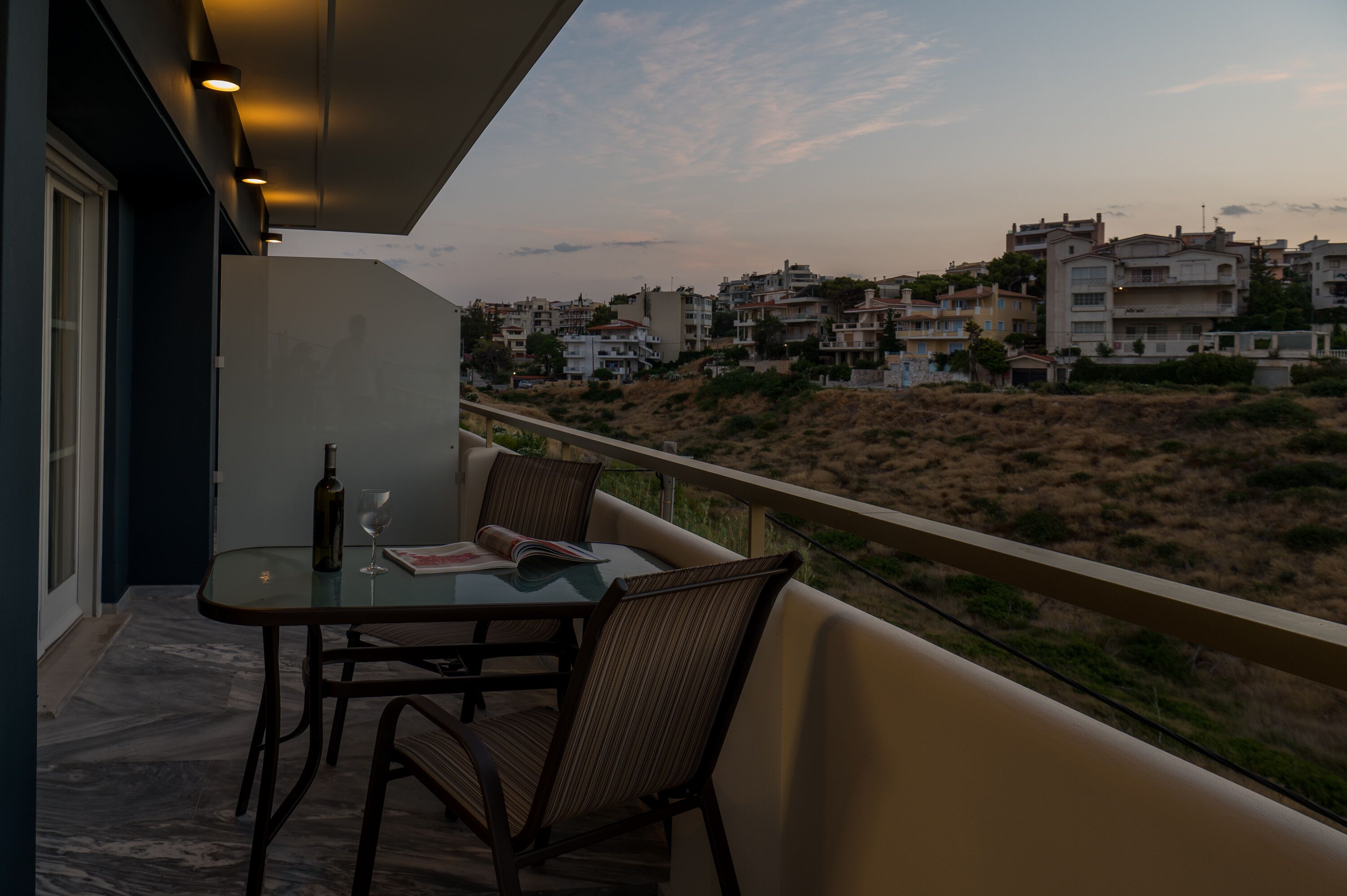 A balcony with a table and chairs, overlooking a residential area with buildings and a hilly landscape.