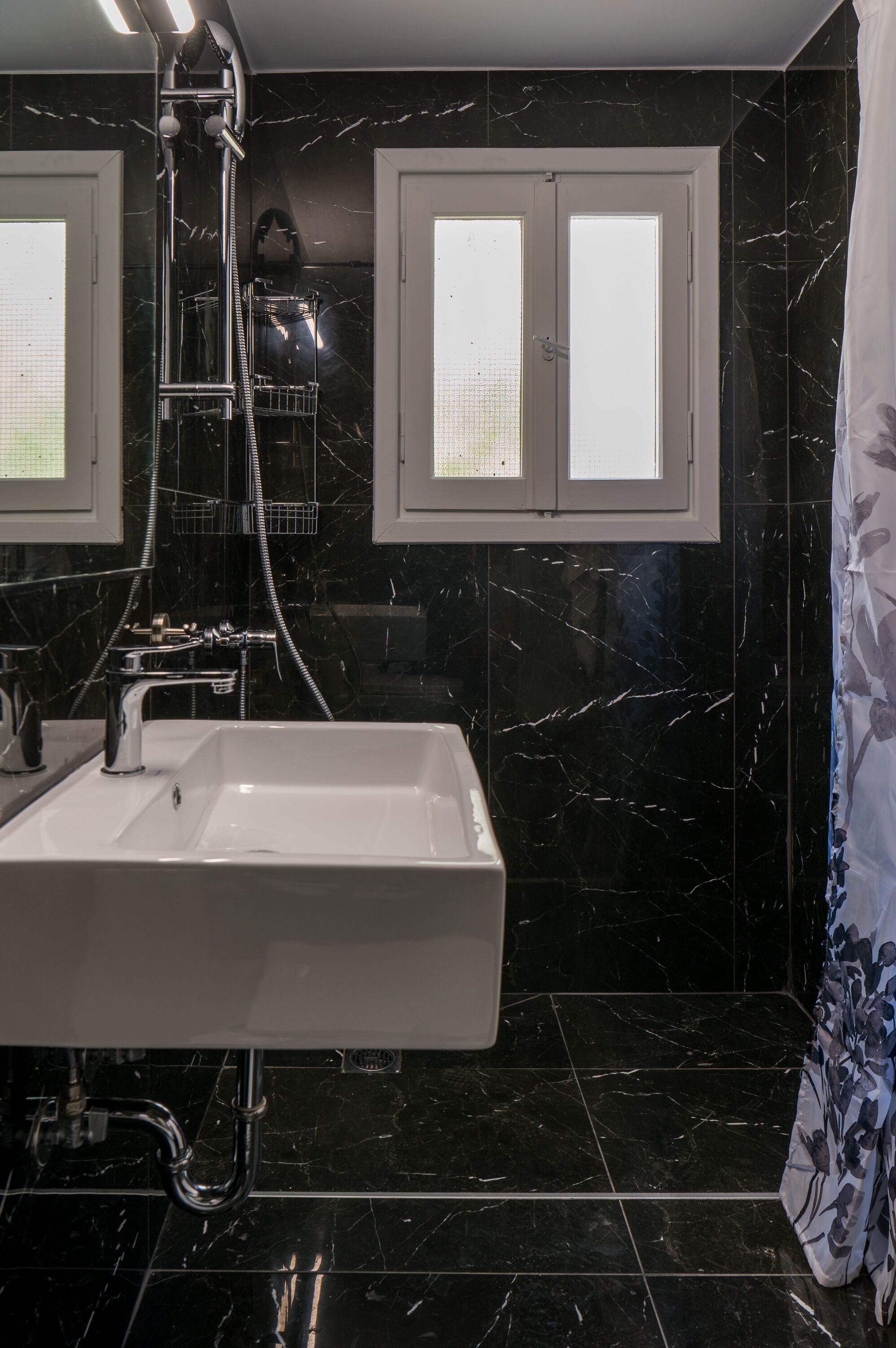 Un baño con lavamanos blanco, paredes de mármol negro y ducha con grifería cromada.