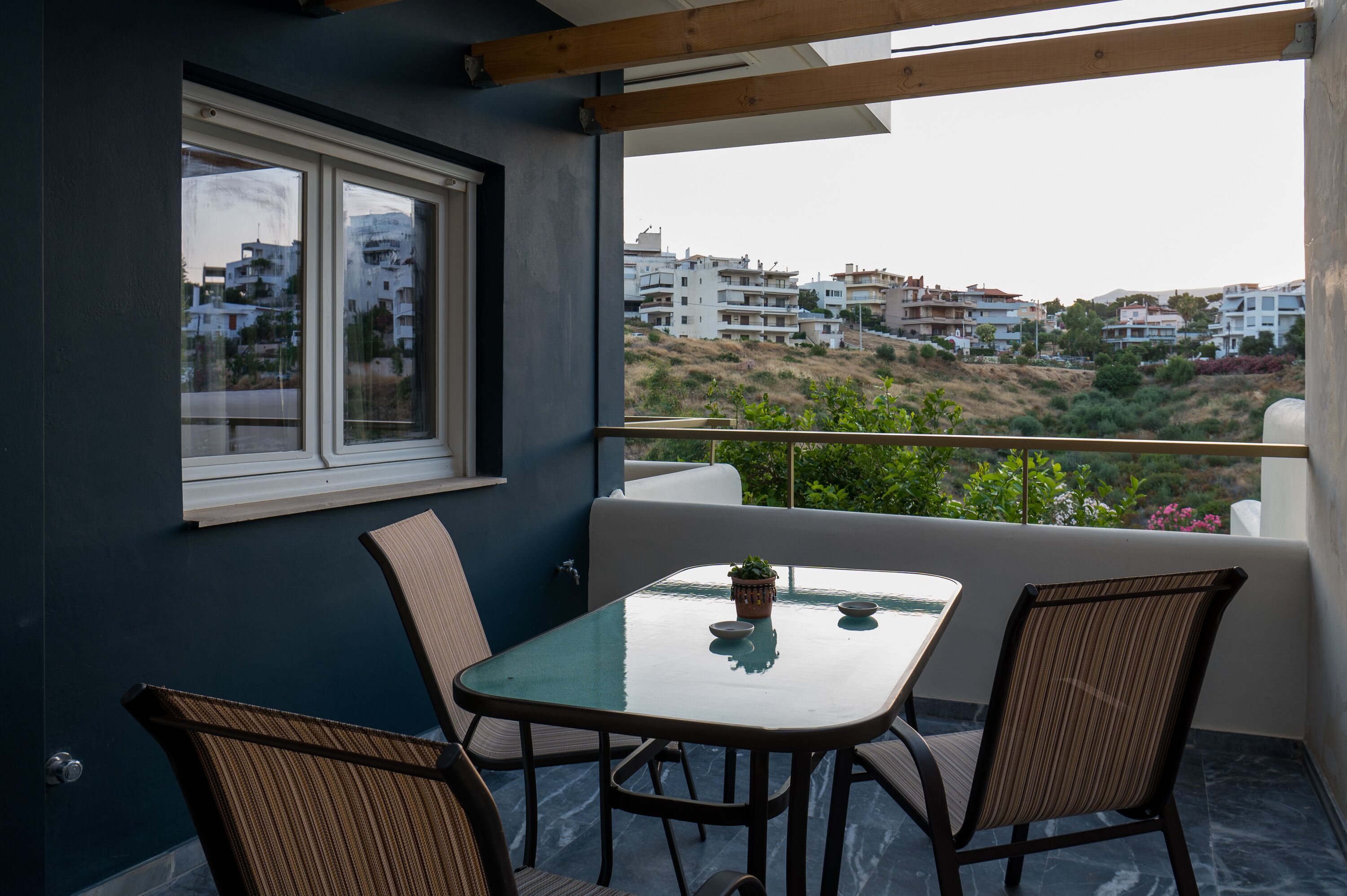 A balcony with a table and chairs, a window, and a view of buildings in the distance.