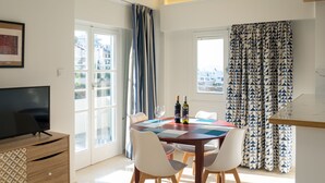A dining area with a table set for four, a television on a wooden cabinet, and a city view through the windows. - CHROMA Lodge Rafina (Rafina-Pikermi)