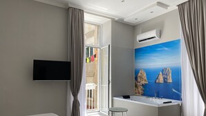 A modern hotel room with a bed, a desk with a sink, a large window with a view of cliffs and the sea, and an air conditioning unit. - Toledo Relax (Naples)