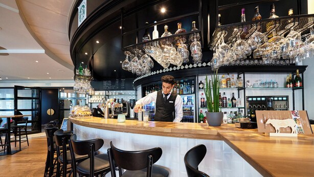 Zona bar con un barista, sgabelli da bar, diverse bottiglie e un lampadario di vetro decorativo.