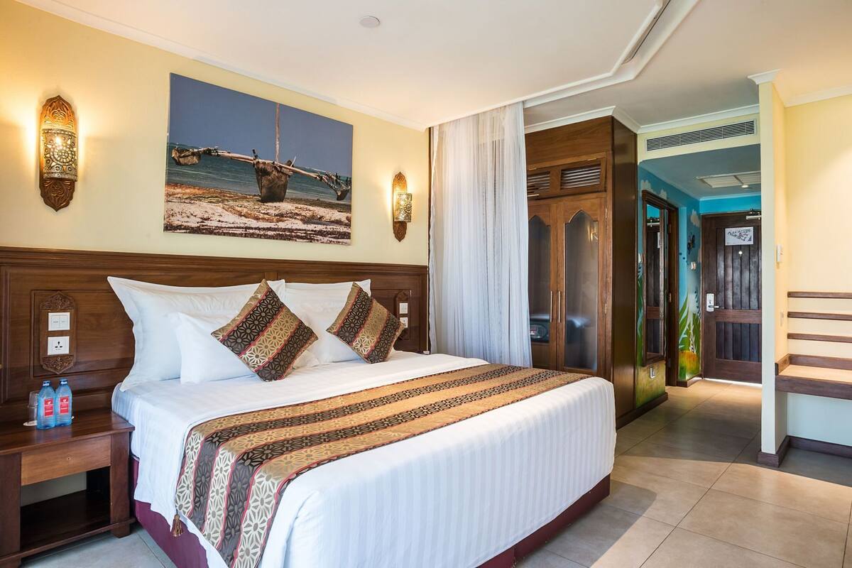 A hotel room with a large bed, a nightstand with water bottles, a painting of a beach scene, and a wooden wardrobe.