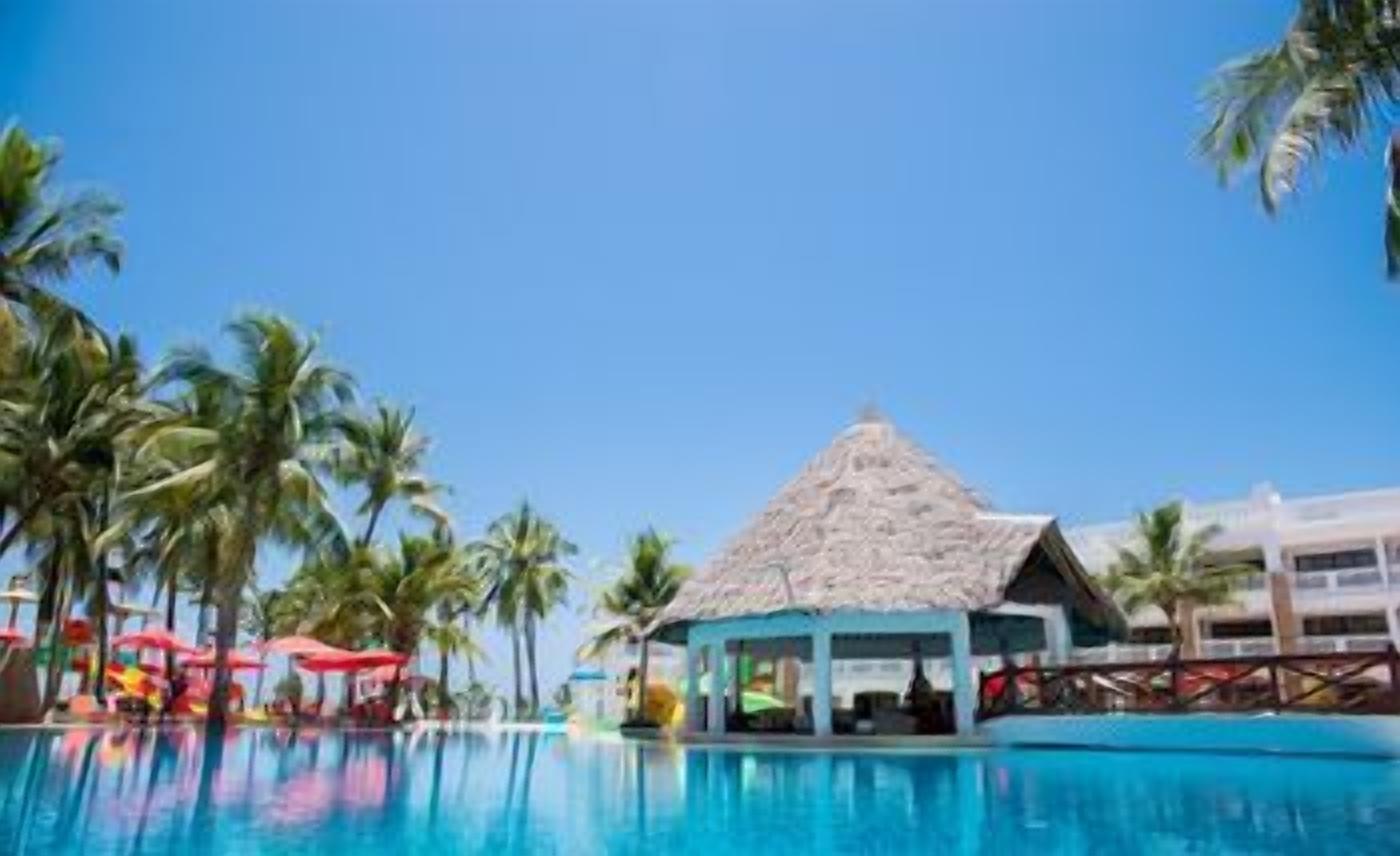 A pool area with a thatched-roof structure, palm trees, and a clear blue sky.