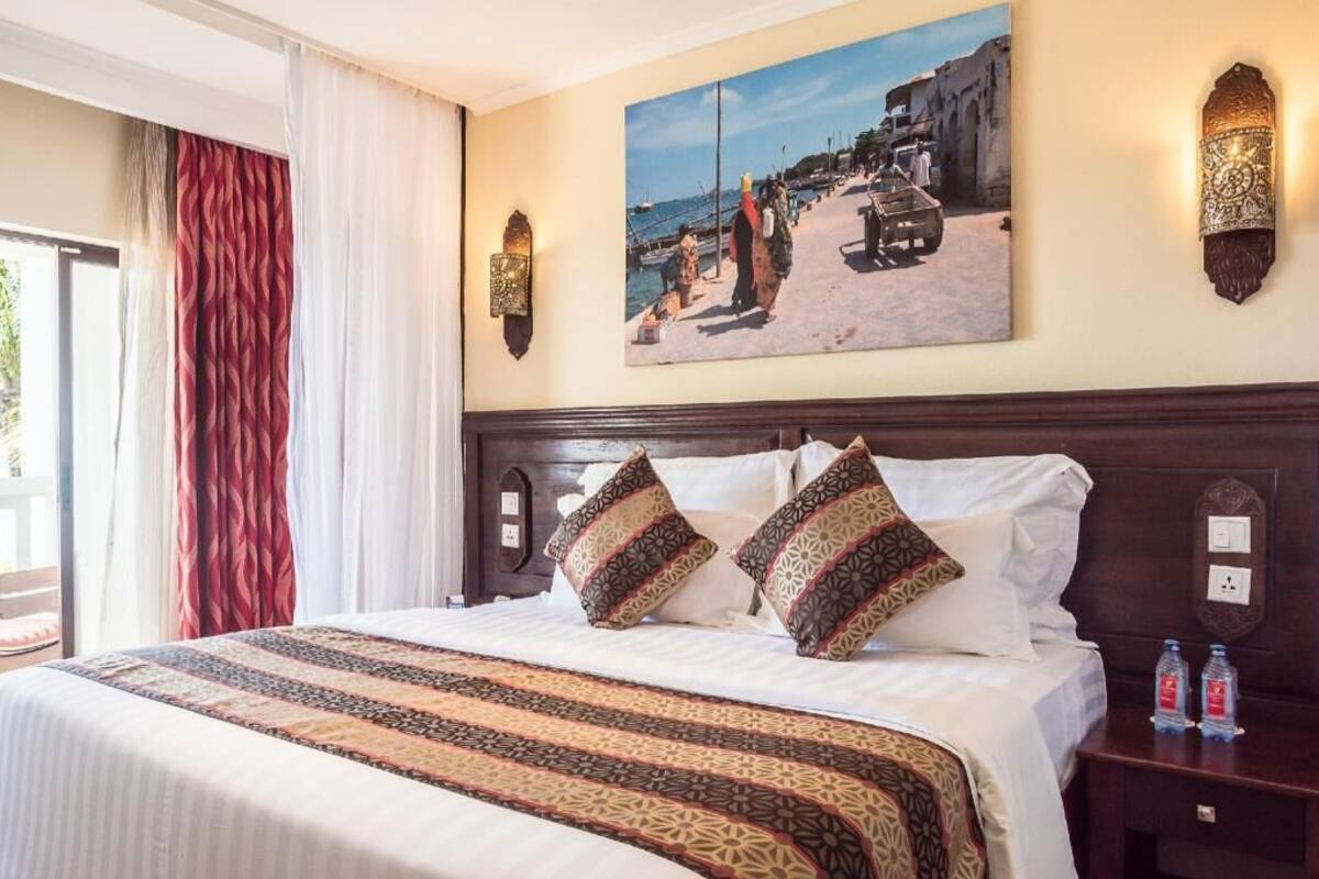 A hotel room with a bed, a painting of a beach scene, a nightstand with water bottles, and a window with curtains.