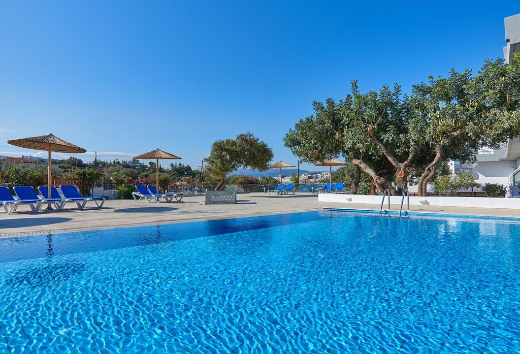 A clear blue pool with lounge chairs and umbrellas, surrounded by trees and buildings.