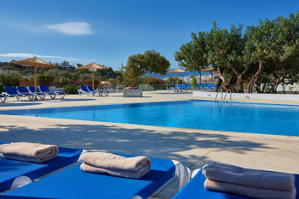 A pool area with lounge chairs and umbrellas, surrounded by trees and a clear sky.
