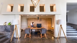 A modern lobby with a desk, two chairs, a computer, and a person working. There are two lamps on stands, a staircase to the right, and a wooden wall with built-in lighting. - Vasia Ormos Hotel (Agios Nikolaos)
