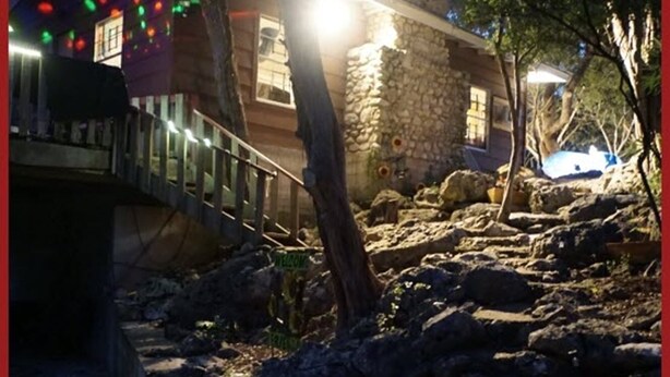 Un chemin de pierres menant à un bâtiment à la façade en pierre et doté d’une terrasse en bois illuminée de guirlandes lumineuses.