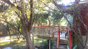 Un pont en bois avec une rambarde rouge menant à un bâtiment rouge, entouré d’arbres et de verdure.