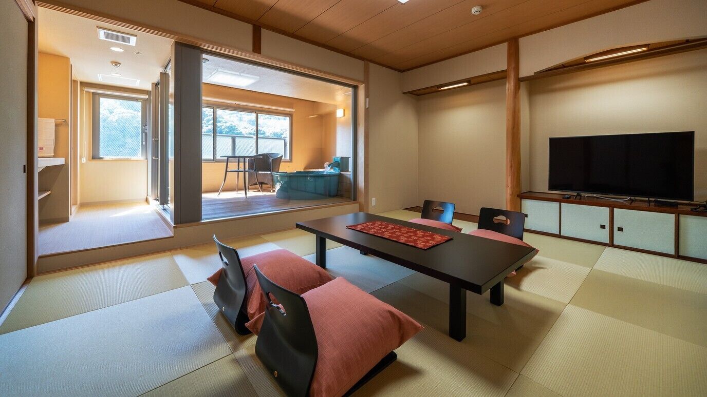 A traditional Japanese-style room with tatami flooring, a low table with cushions, a television, and a view of the outdoors.