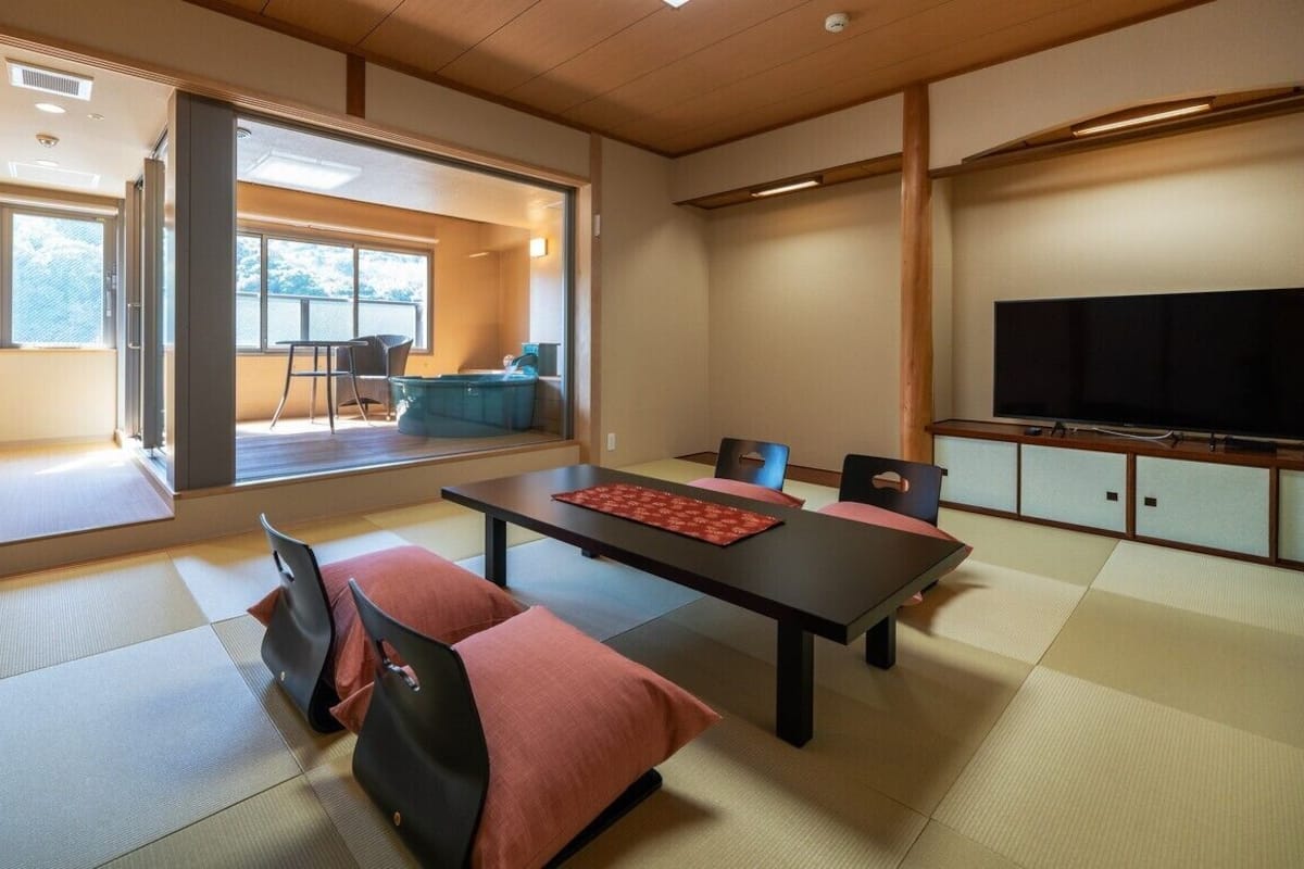 Una habitación tradicional japonesa con suelo de tatami, una mesa baja con cojines, televisión y vistas al exterior.