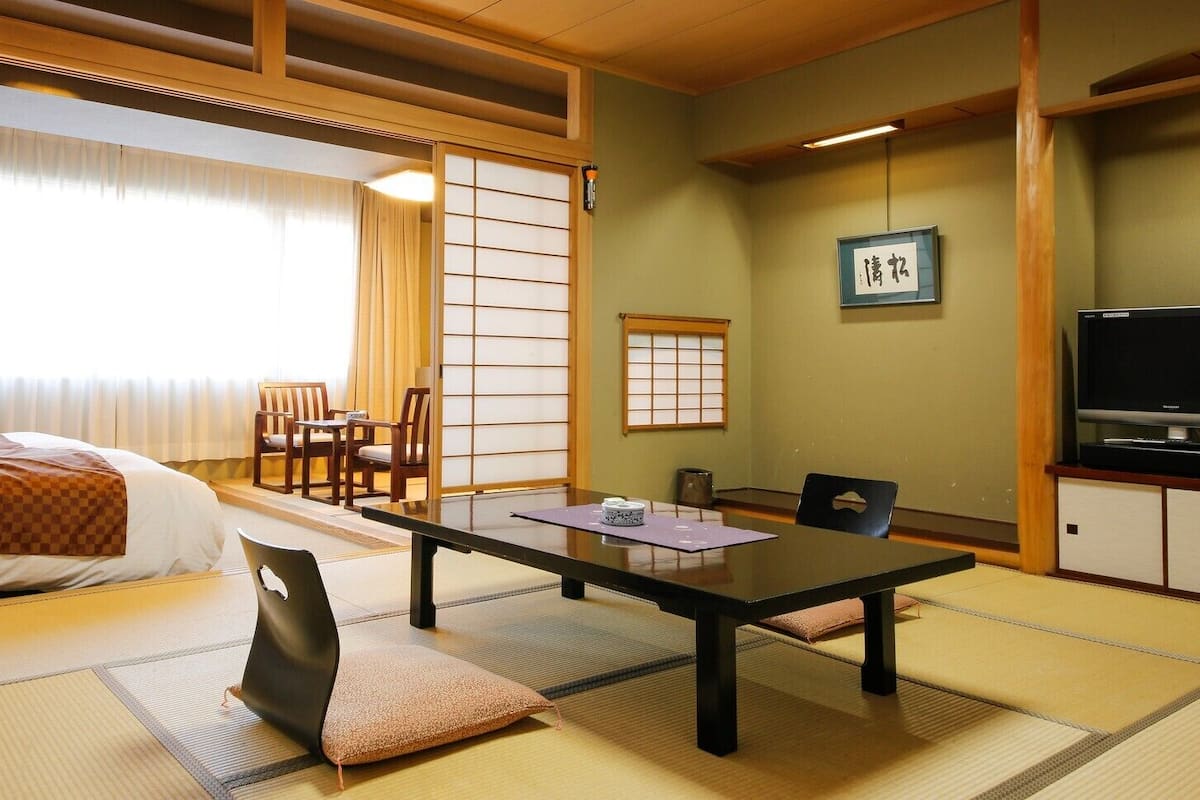 Una habitación tradicional japonesa con suelo de tatami, una cama, una mesa con cojín y un televisor.