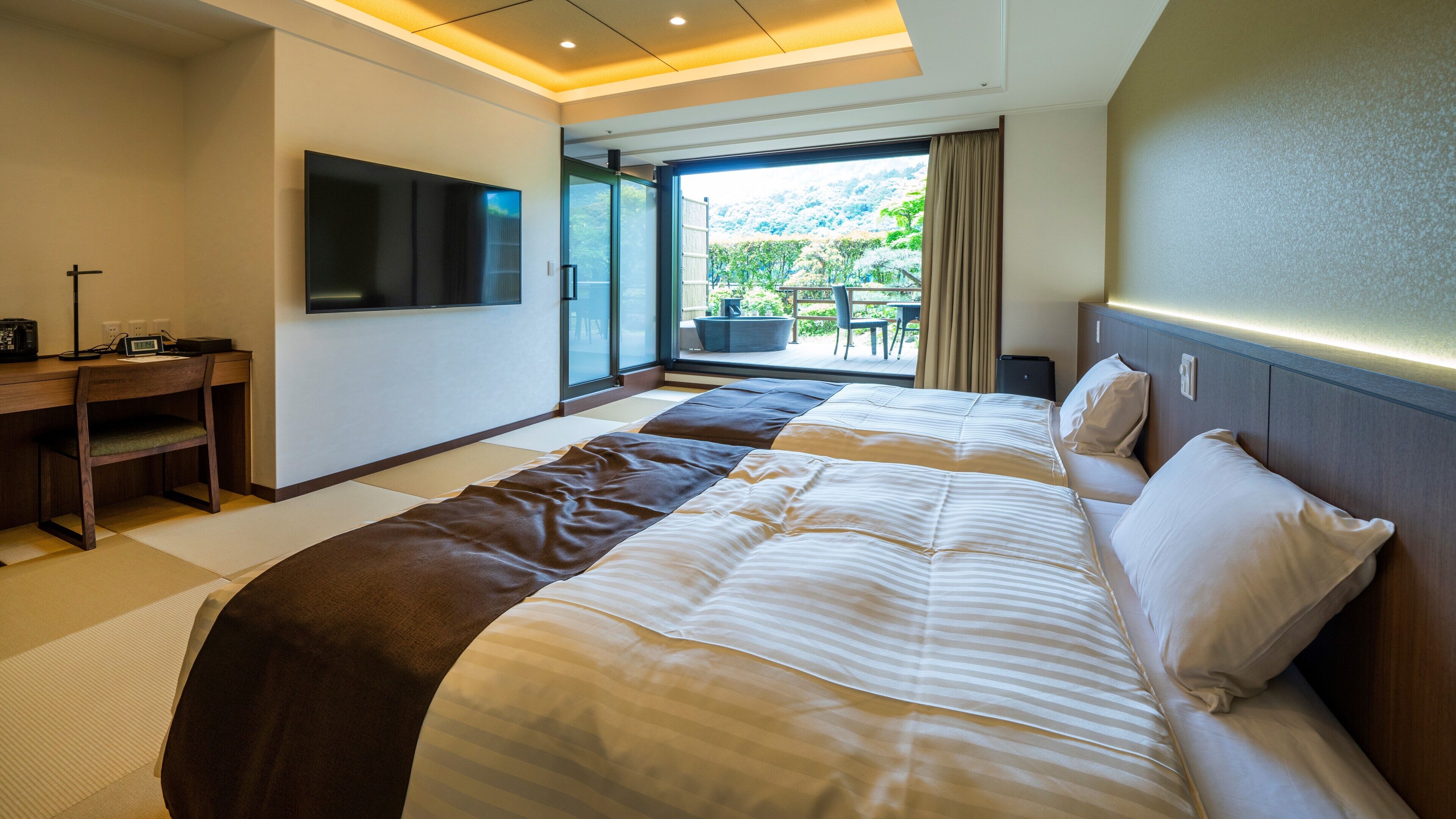 A hotel room with a large bed, a desk with a flat-screen TV, and a view of a garden through a sliding door.