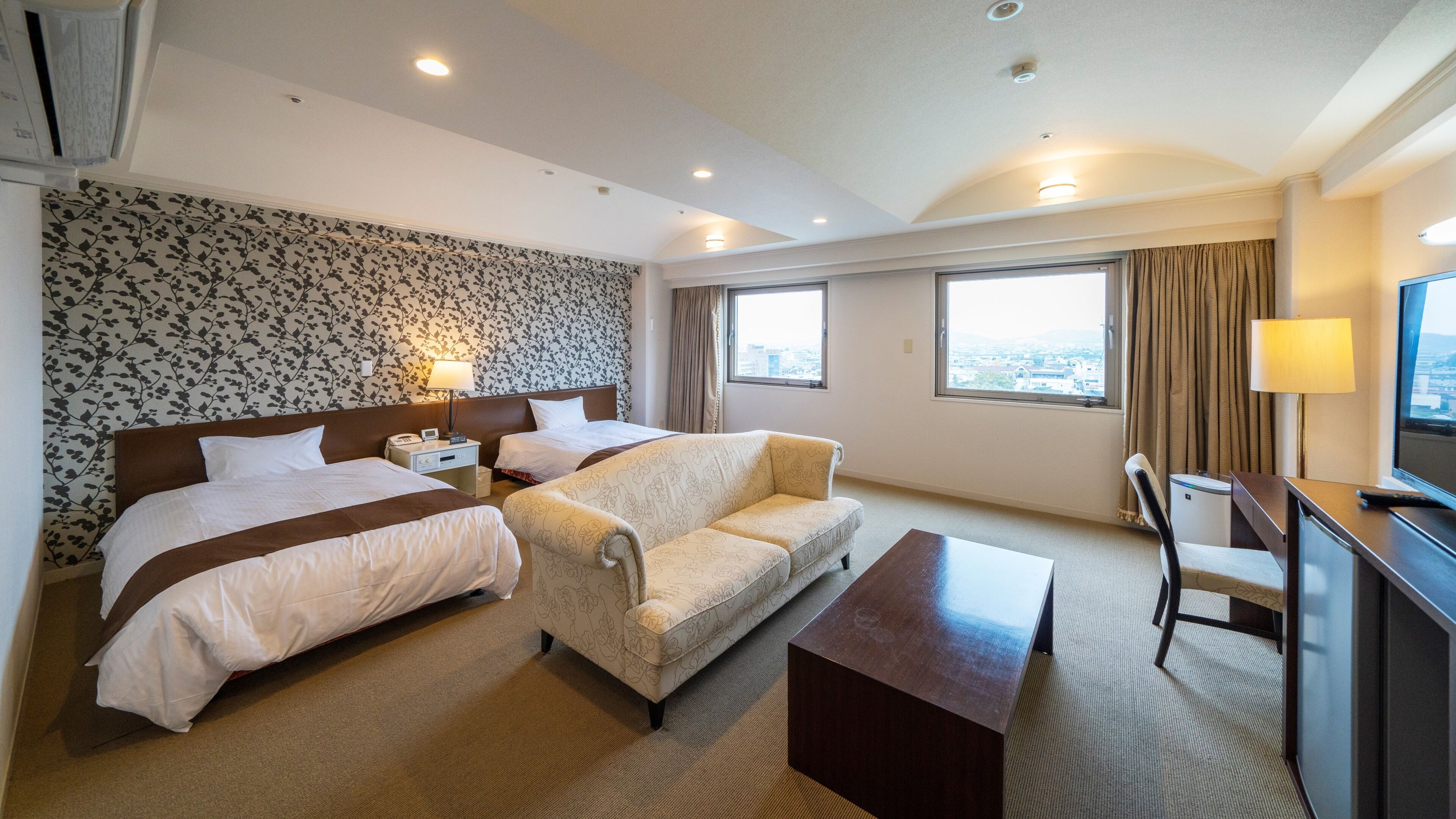 A hotel room with two beds, a sofa, a desk, and a view of the city.