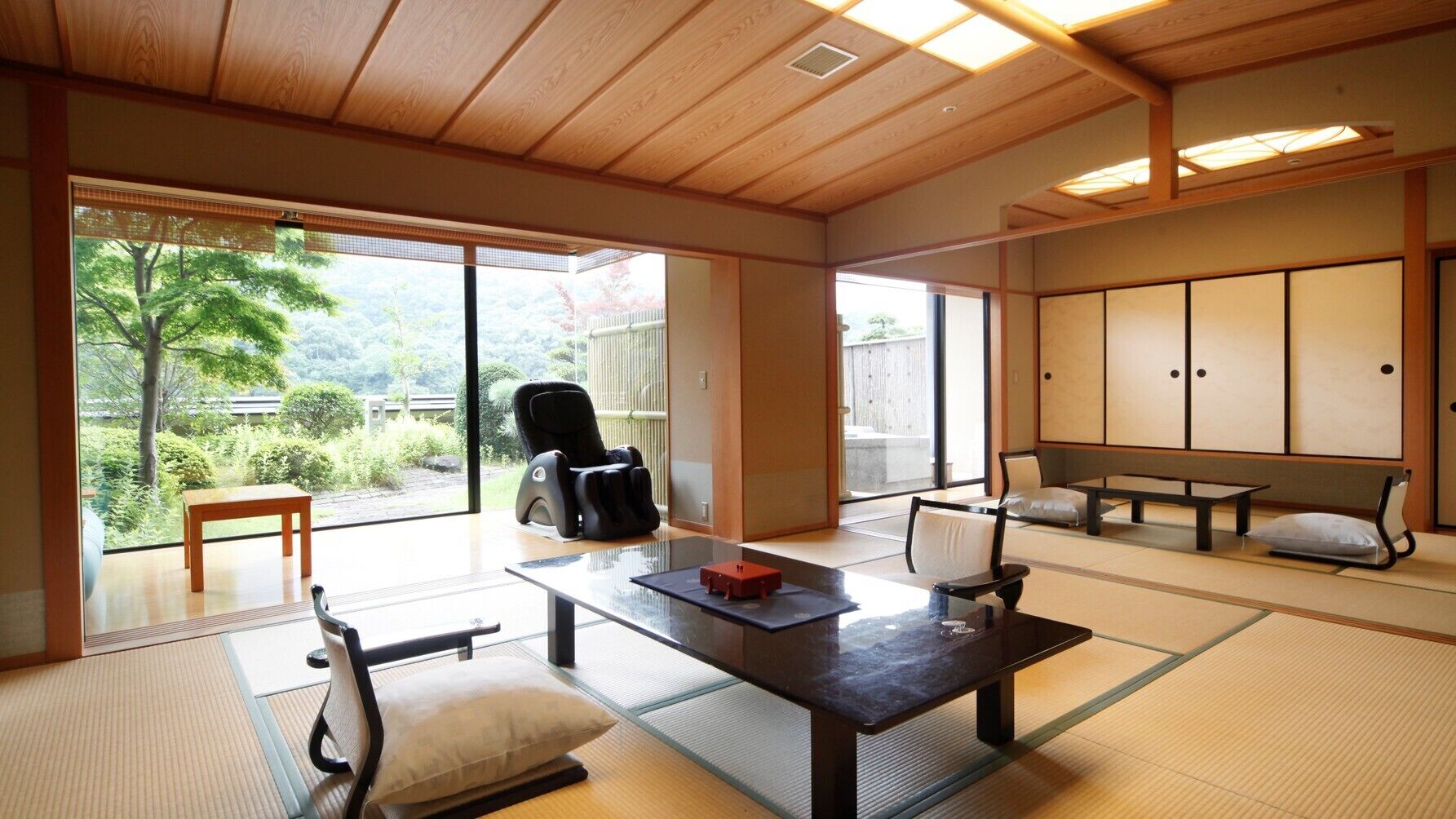 A traditional Japanese room with tatami flooring, a tatami mat table, a black massage chair, a wooden bench, and sliding doors leading to a garden.