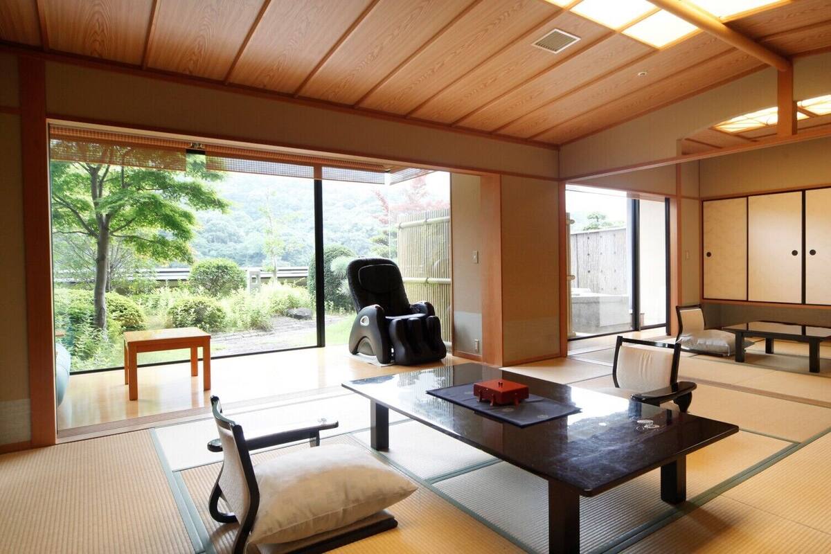 Una habitación japonesa tradicional con suelo de tatami, una mesa baja de tatami, una silla de masaje negra, un banco de madera y puertas correderas que dan a un jardín.