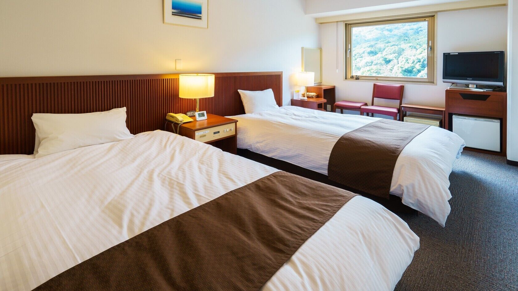 A hotel room with two beds, a desk with a lamp, a chair, a television, and a window with a view.