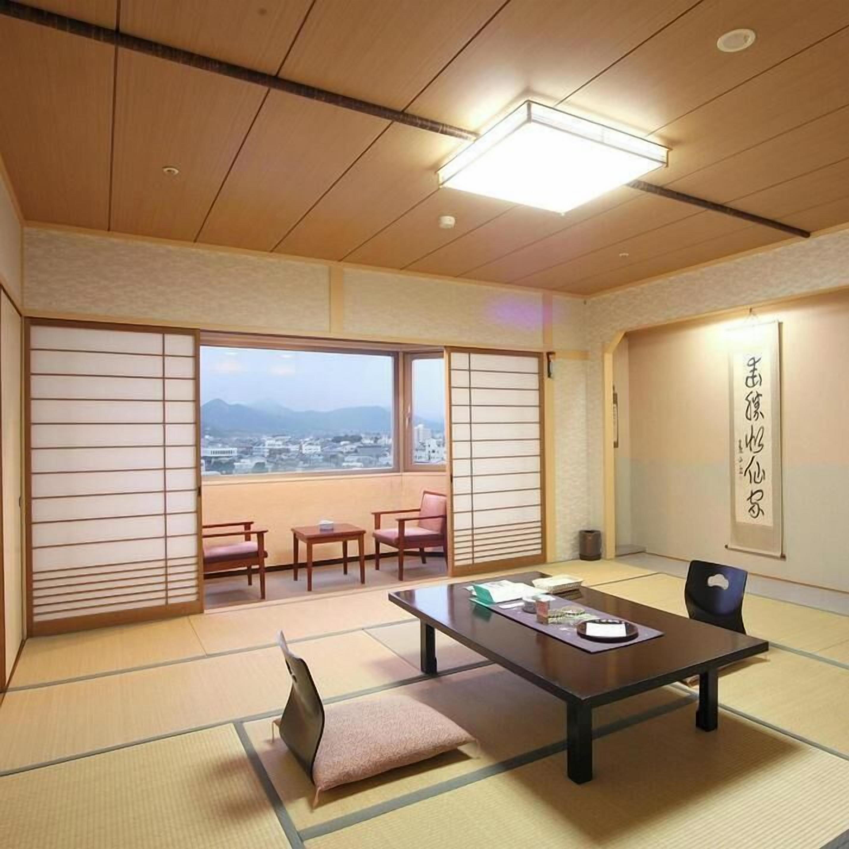 Una habitación japonesa tradicional con suelo de tatami, una mesa baja, sillas y vistas a un paisaje urbano a través de grandes ventanales.