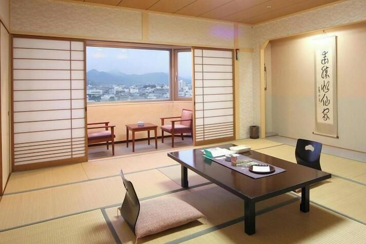 Una habitación japonesa tradicional con suelo de tatami, una mesa baja, sillas y vistas a un paisaje urbano a través de grandes ventanales.