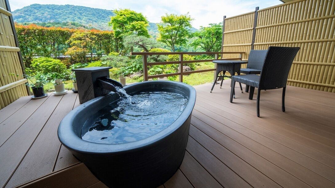Un jacuzzi en una terraza de madera con valla de bambú y sillas, con vista a un jardín con árboles y colinas.