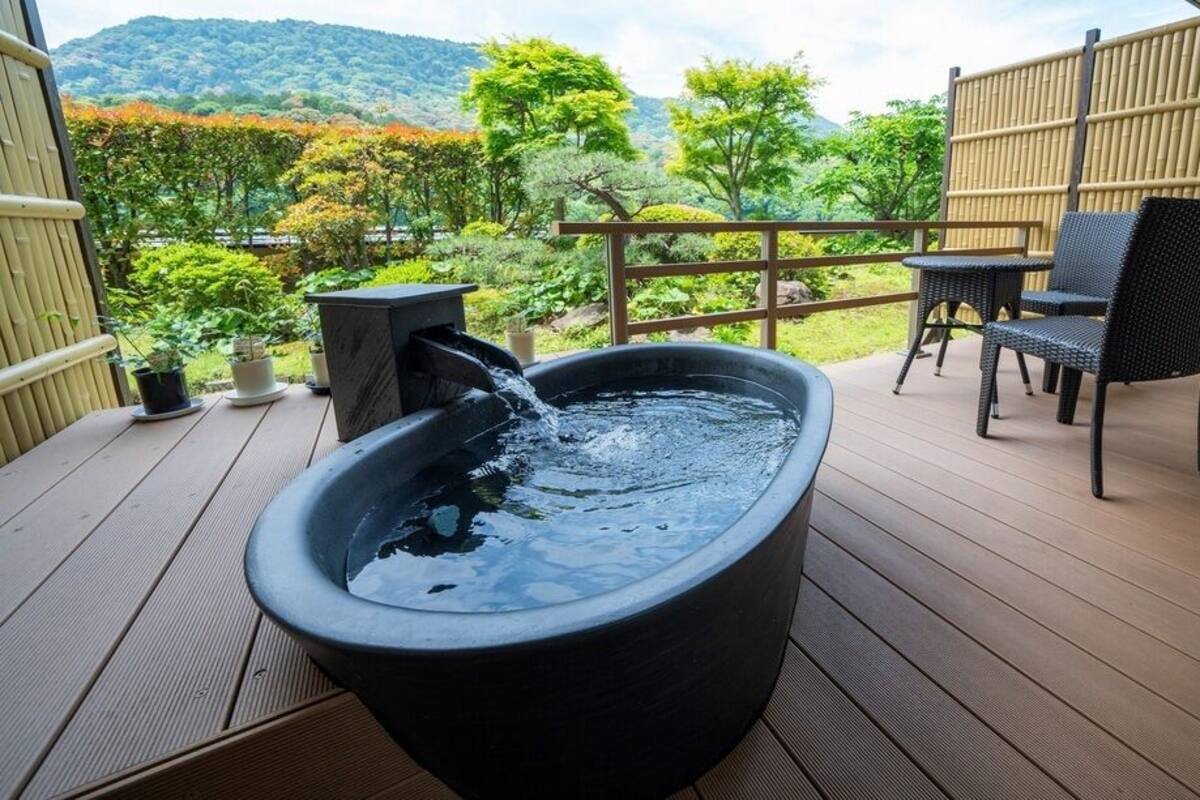 Un jacuzzi en una terraza de madera con valla de bambú y sillas, con vista a un jardín con árboles y colinas.