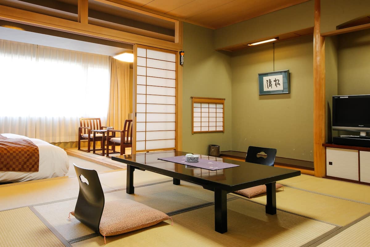 Una habitación de hotel con cama, mesa de comedor, sillas, esteras de tatami, televisor y un biombos shoji.