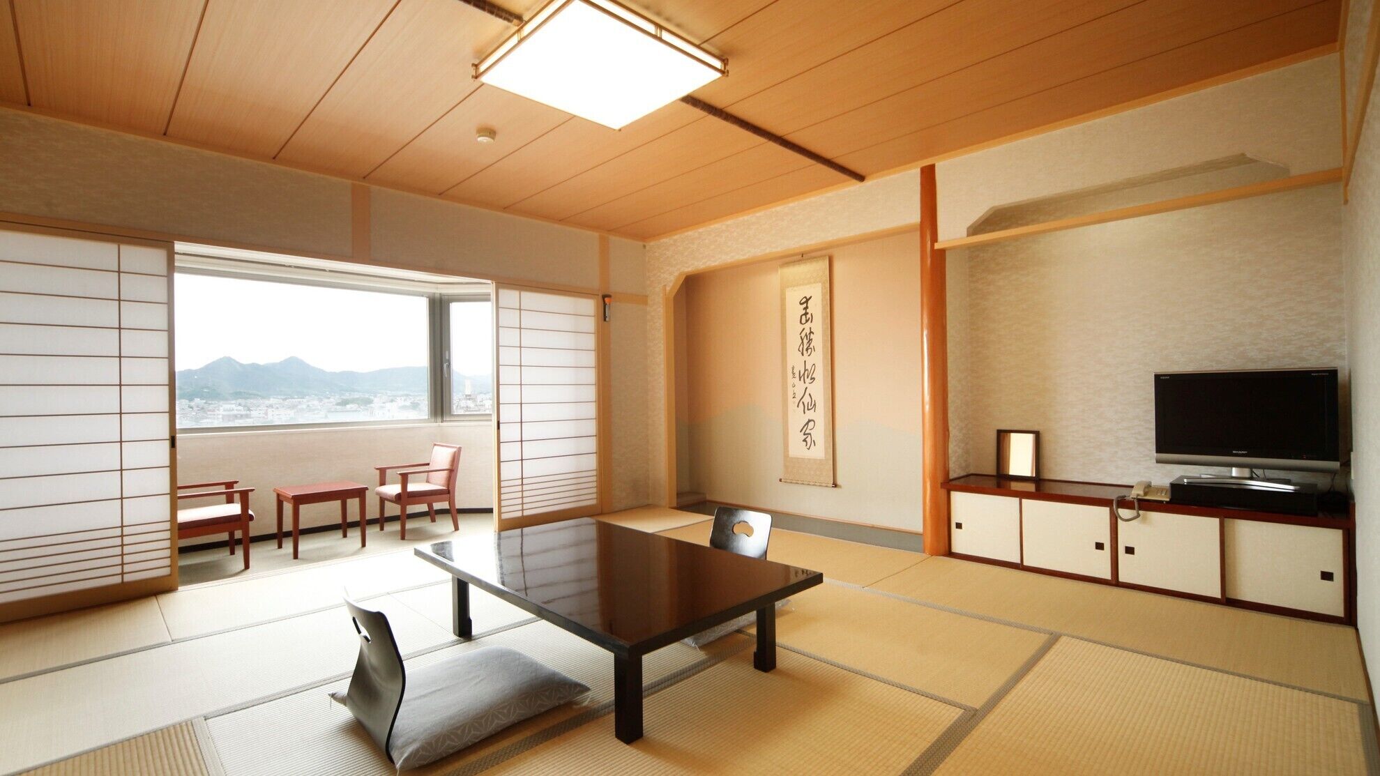 A traditional Japanese room with tatami flooring, a low table, a sofa, a TV, and a view of the mountains.