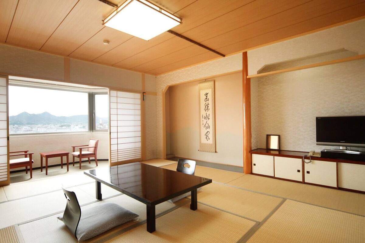 Una habitación tradicional japonesa con suelo de tatami, una mesa baja, un sofá, un televisor y vistas a la montaña.