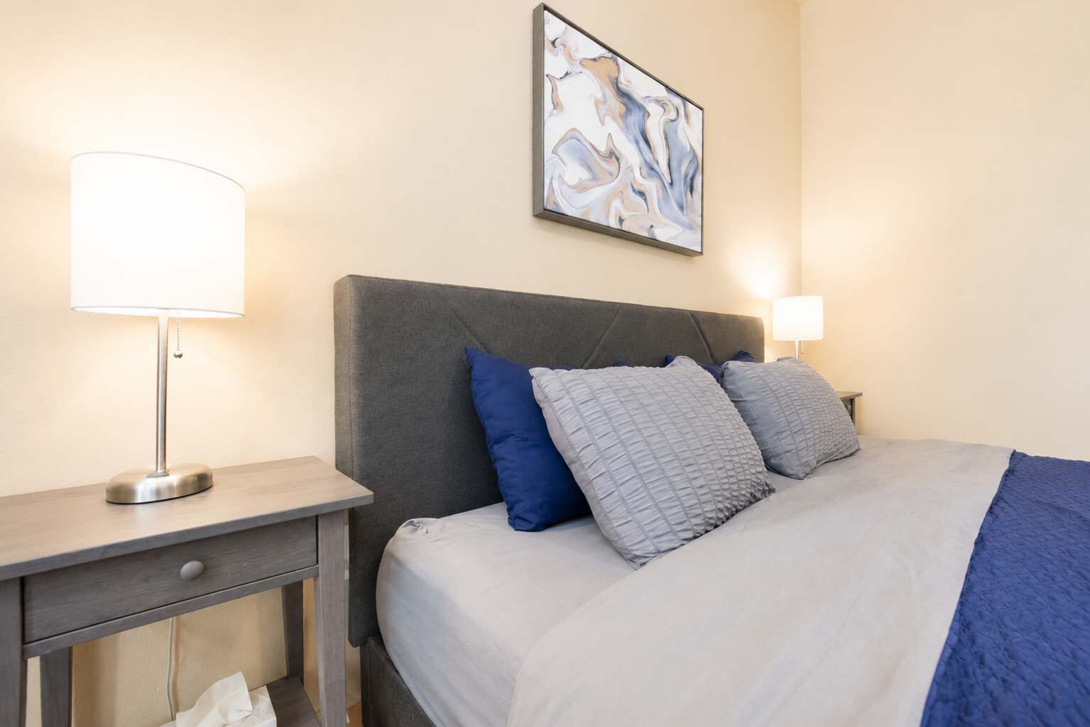Un lit bien fait, avec une tête de lit grise, une table de chevet sur laquelle se trouve une lampe, et un tableau abstrait accroché au mur.