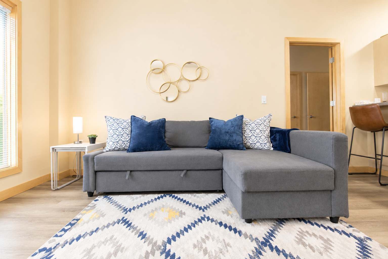 Un sofá seccional gris con cojines estampados en azul y blanco, una mesita auxiliar blanca con una lámpara y un adorno en la pared.
