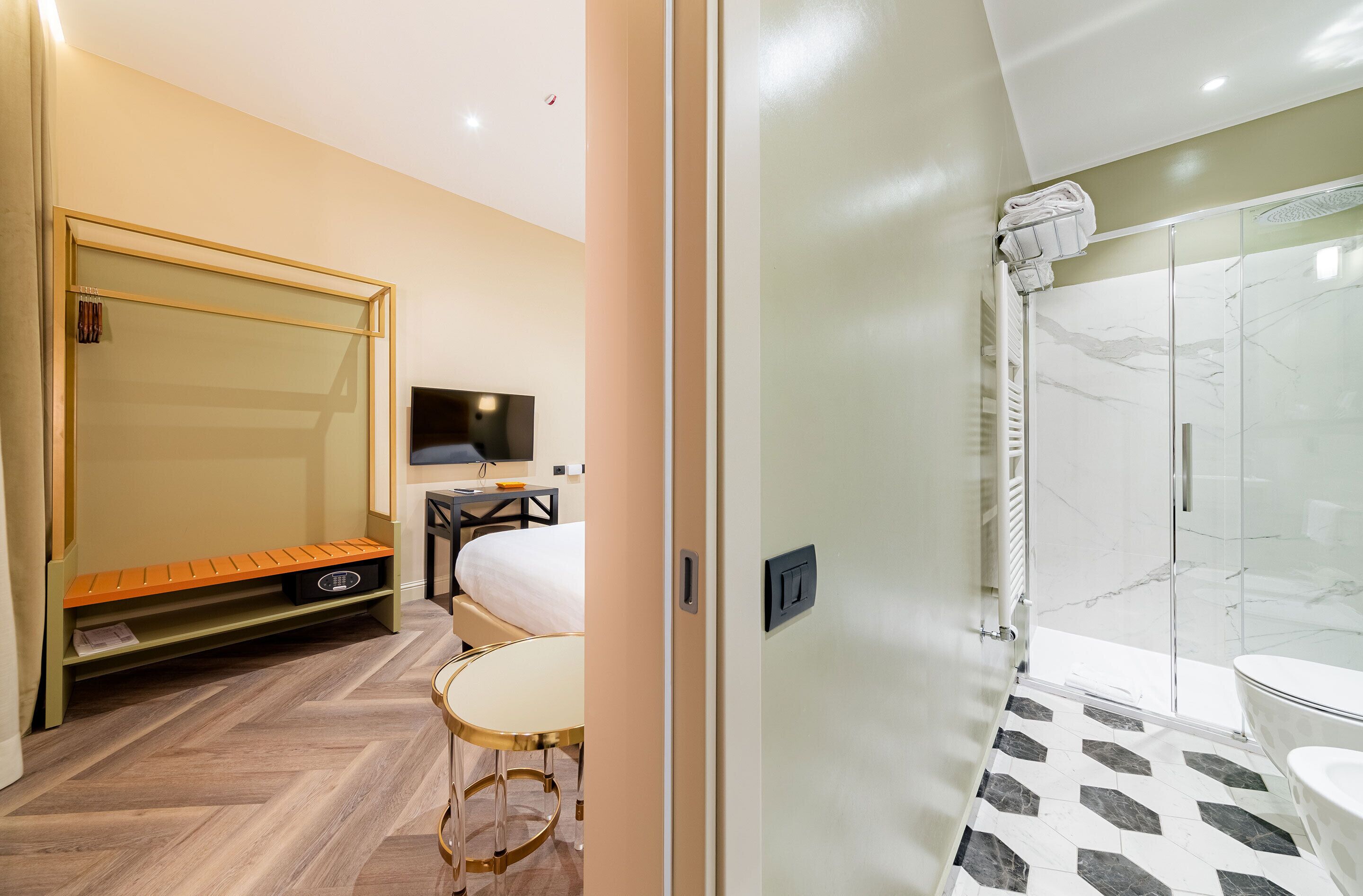 A hotel room with a bed, a bench, a small table, a TV, and a shower area with a glass door.