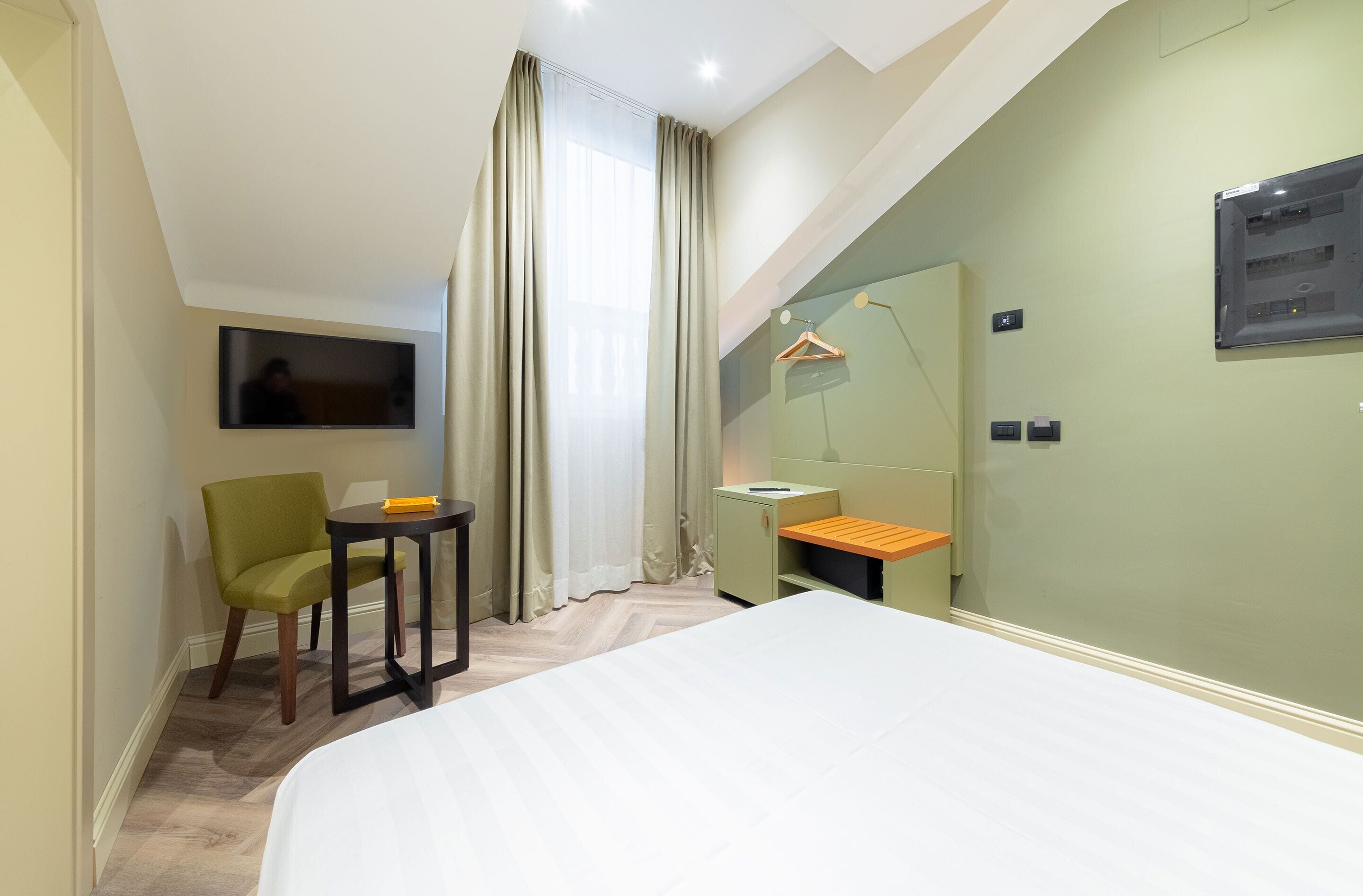 A hotel room with a bed, a desk, a chair, a TV, a mirror, and a window with curtains.