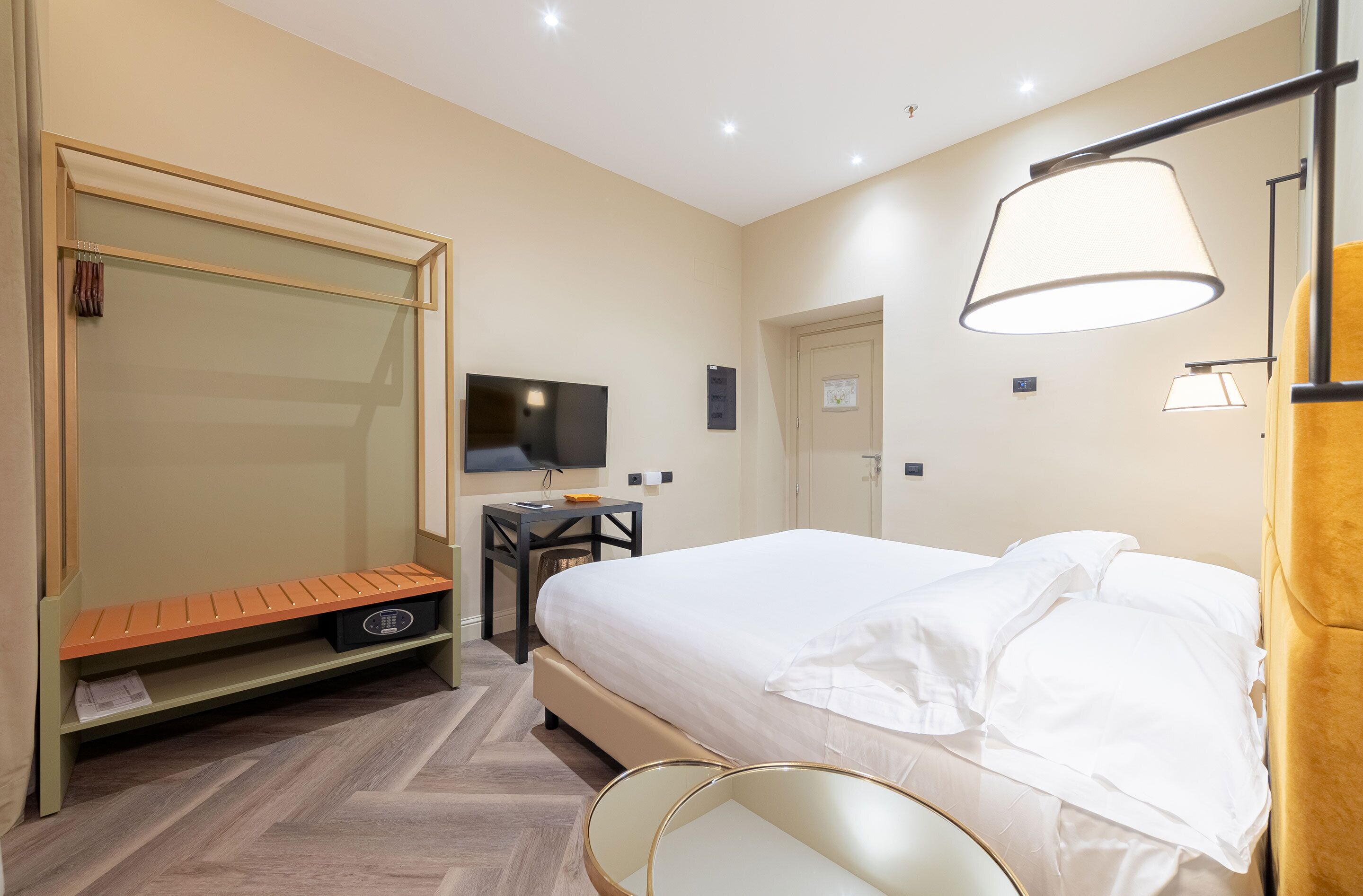 A hotel room with a bed, a desk with a TV, a wardrobe, and a bench.