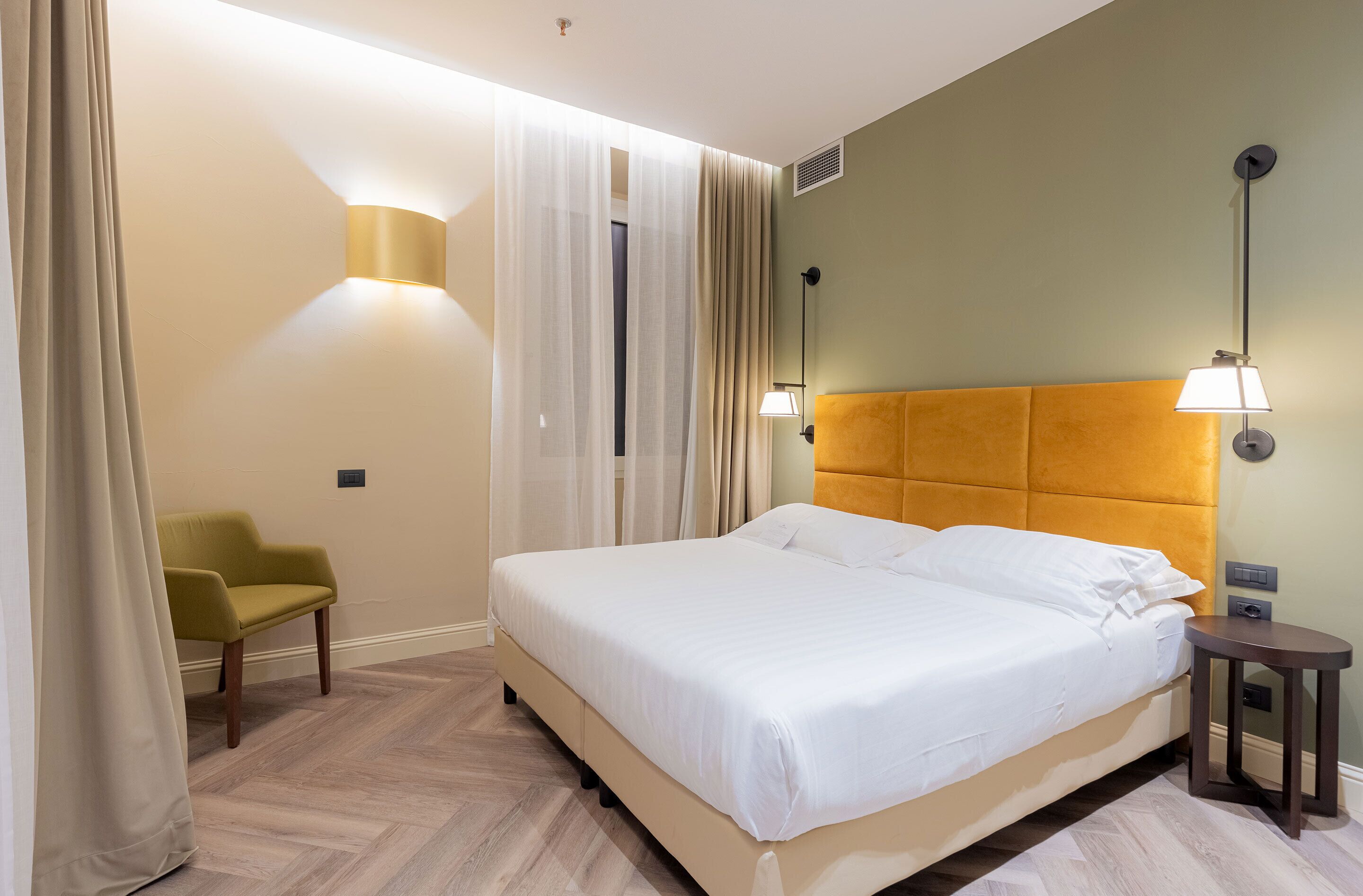 Quarto de hotel bem arrumado, com uma cama grande, uma cadeira pequena e duas luminárias fixadas na parede.