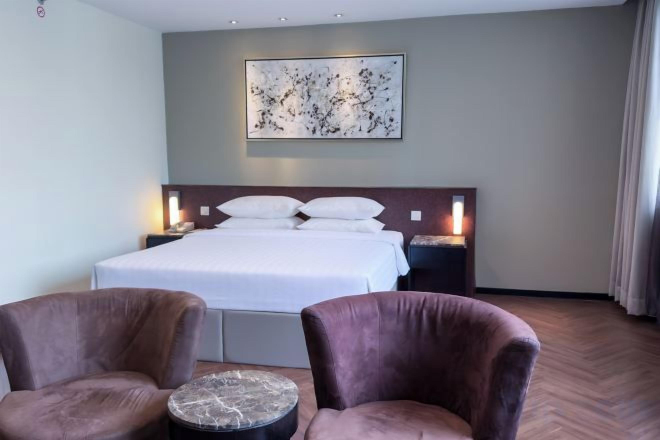 A hotel room with a large bed, two armchairs, a small round table, a nightstand with a lamp, and a painting on the wall.