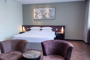 Une chambre d’hôtel avec un grand lit, deux fauteuils, une petite table ronde, une table de chevet avec une lampe et un tableau au mur.