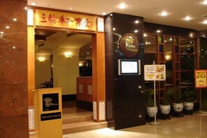 Entrada de hotel con mostrador de recepción, letrero y plantas en macetas.