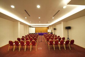 Un salón de conferencias con filas de sillas rojas, un atril, una pantalla y un orador.