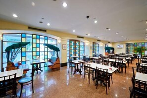 A spacious indoor dining area with multiple tables and chairs, large windows with view-enhancing curtains, and a ceiling with recessed lighting.