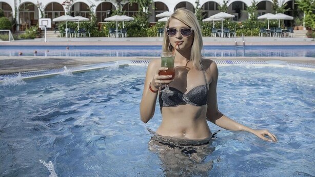 A person in a swimming pool with a drink, surrounded by outdoor seating and a building with arches. - Irene Palace (Rhodes)