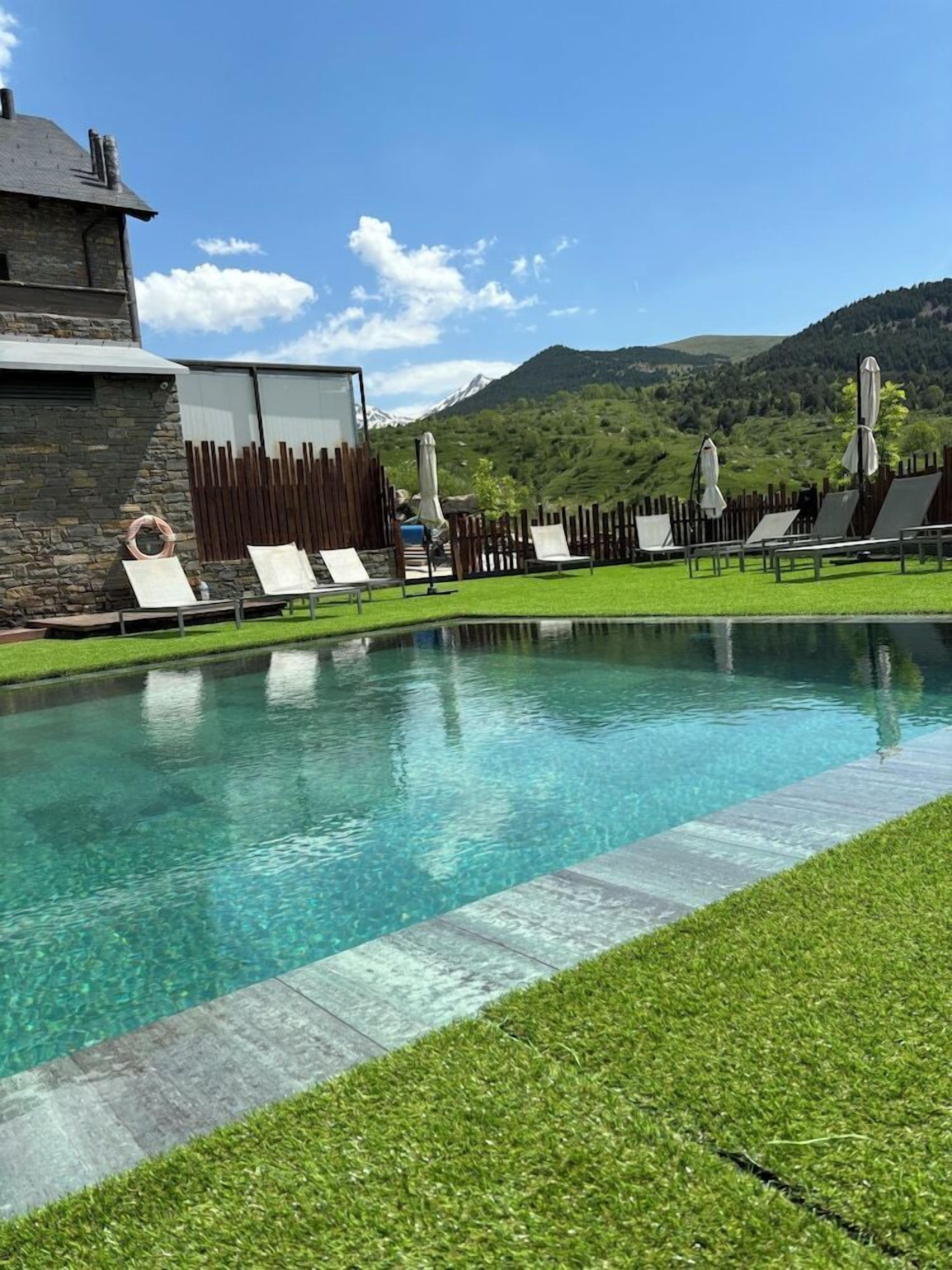 Una piscina con sillones y sombrillas, rodeada por una valla de madera y un edificio de piedra.