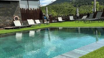 Una piscina con sillones y sombrillas, rodeada por una valla de madera y un edificio de piedra.