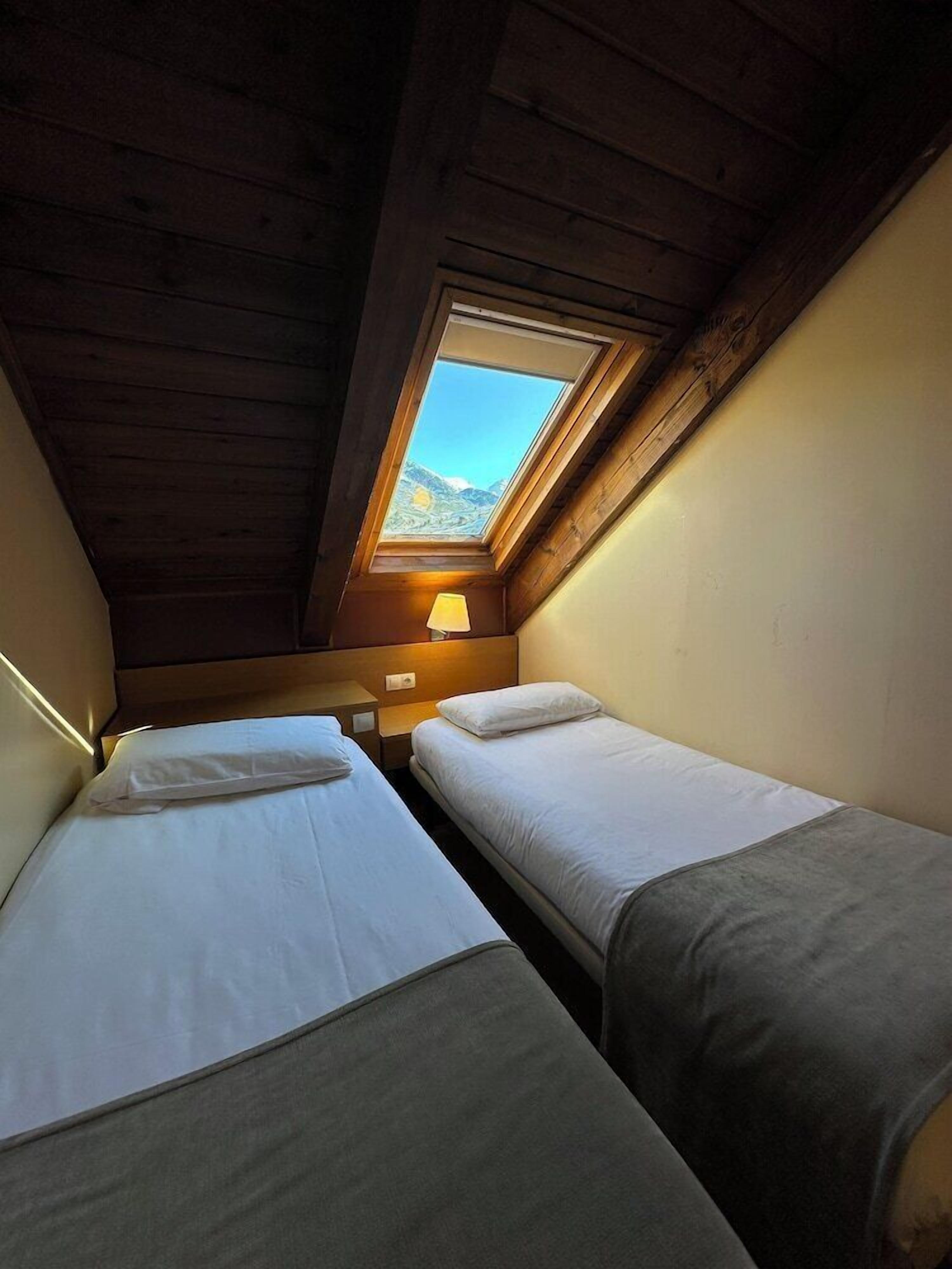A small, cozy room with two single beds, a skylight, and a nightstand with a lamp.