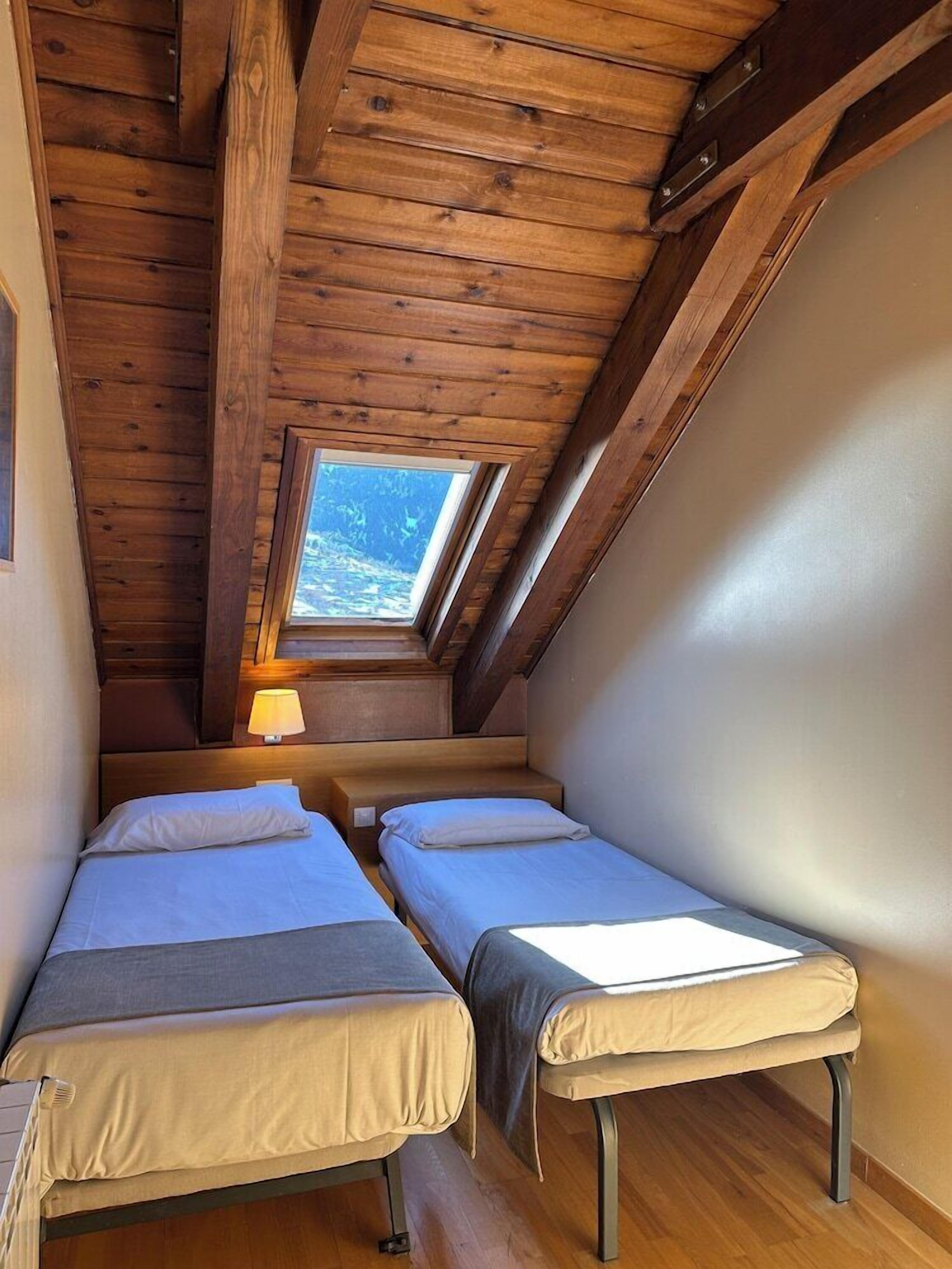 A small, cozy room with two beds, wooden ceiling beams, a skylight, and a view of the outdoors.