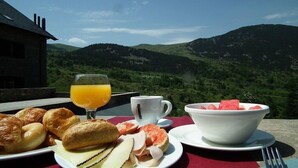 Une table dressée pour le petit-déjeuner avec des croissants, un bol de pastèque, un verre de jus d’orange et une tasse, le tout offrant une vue sur un paysage montagneux.